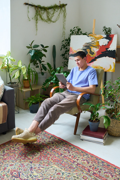 A guy with dark hair dressed in a T-shirt and corduroy trousers uses a tablet sitting in an armchair with wooden legs and armrests in a room with brightly painted canvas on an easel with green plants and a Turkish carpet